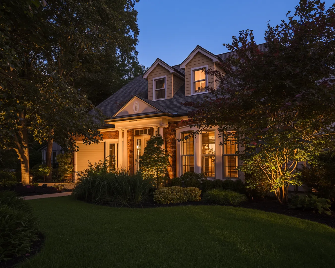 House exterior with illuminated porch, trees and landscape at dusk